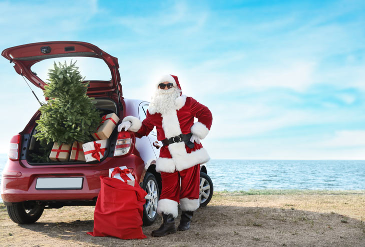 On a beach, a red car with a Christmas tree and presents and santa stood next to it.
